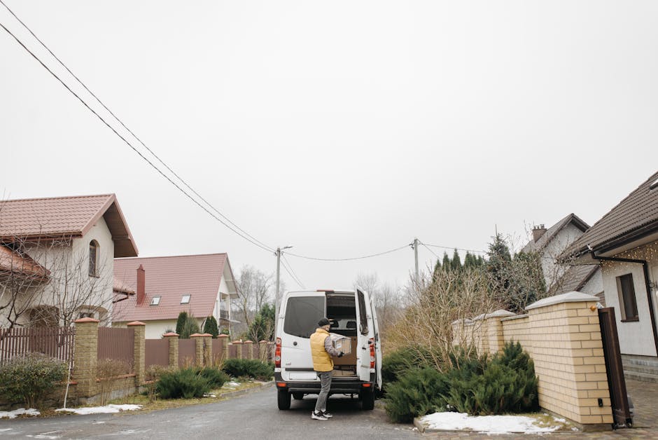 A person loading a cardboard box into the back of a white moving van parked along a narrow residential street with detached houses featuring pitched roofs, brick and rendered walls, and front gardens.